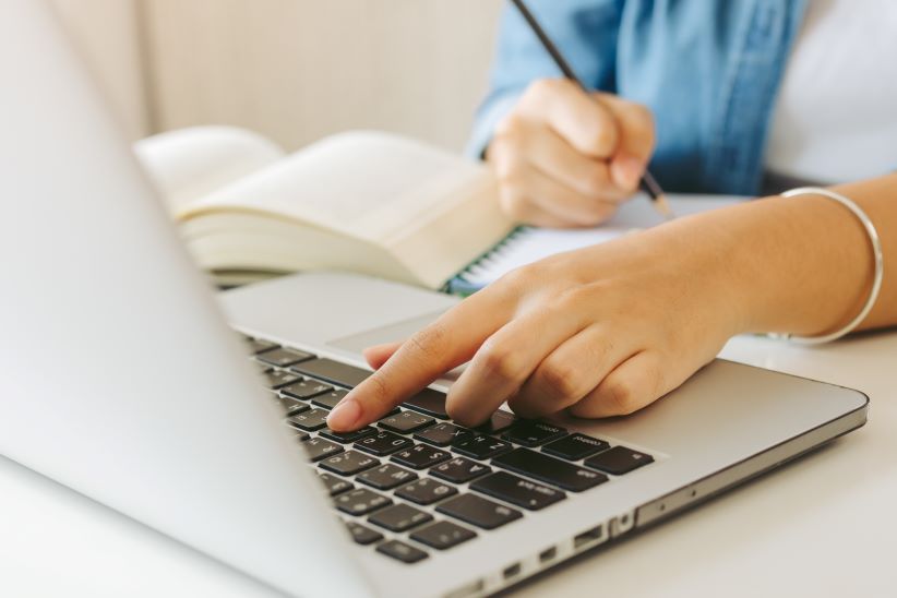 Egy asztalra fektetett laptop és a rajta dolgozó női kéz látszik egy könyvvel és egy tollal jegyzetelő kézzel 