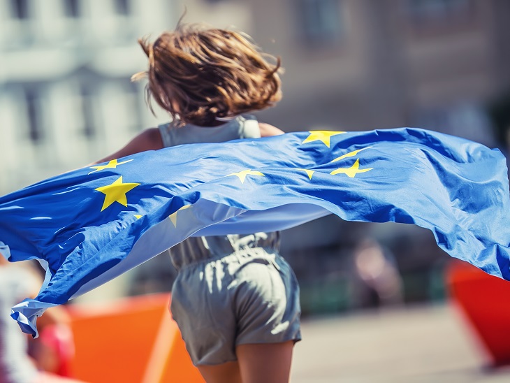 Barna hajú kisgyerek szalad egy EUs zászlóval a hátán