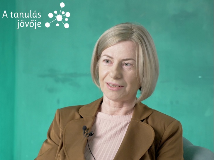 Szőke hölgy barna zakóban zöld háttér előtt interjút ad