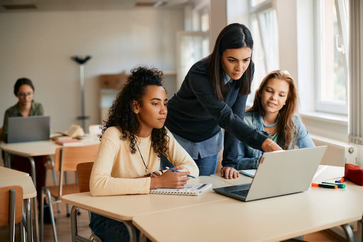 Fiatal lányok ülnek egy laptop előtt és egy idősebb társuk segít nekik