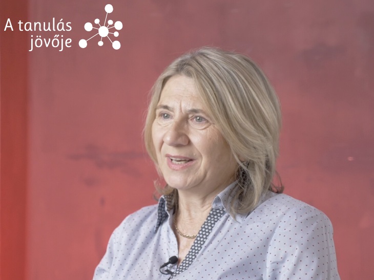 Szőke hölgy ingben interjút ad piros háttér előtt